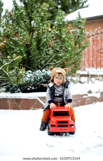 Cute baby boy in cozy winter\
clothes and funny hat playing with toy car in snow on beauty winter\
day