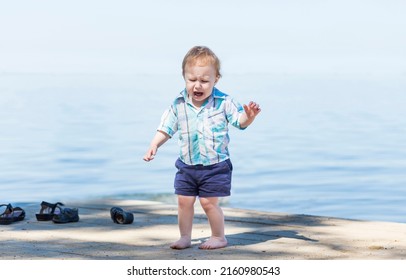 Cute Baby Boy Is Crying On The Beach With A Beautiful Blue Sea In The Background. Portrait Of An Alone Sad Child On A Beach In A Sunny Day.