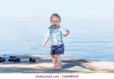 Cute Baby Boy Is Crying On The Beach With A Beautiful Blue Sea In The Background. Portrait Of An Alone Sad Child On A Beach In A Sunny Day.