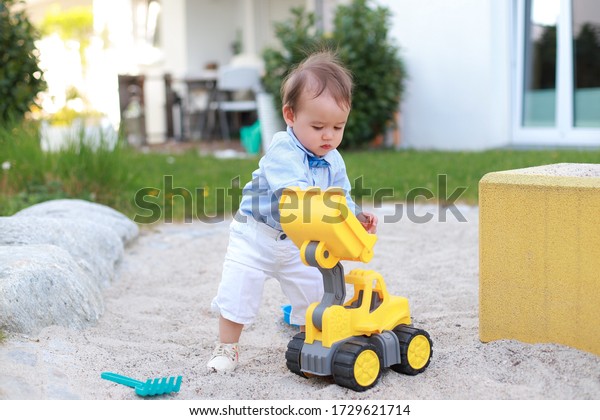 Cute\
baby boy first standing while playing with excavator toy on sand at\
playground.mixed race Asian-German infant wearing blue shirt white\
pants and shoes fashion kid summer outdoor\
park.