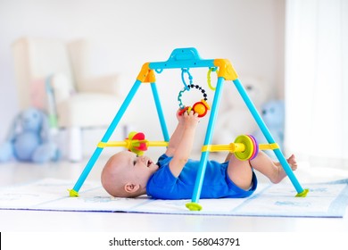 Cute baby boy on colorful playmat and gym, playing with hanging rattle toys. Kids activity and play center for early infant development. Newborn child kicking and grabbing toy in white sunny nursery.