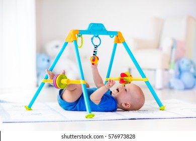 Cute Baby Boy On Colorful Playmat And Gym, Playing With Hanging Rattle Toys. Kids Activity And Play Center For Early Infant Development. Newborn Child Kicking And Grabbing Toy In White Sunny Nursery.