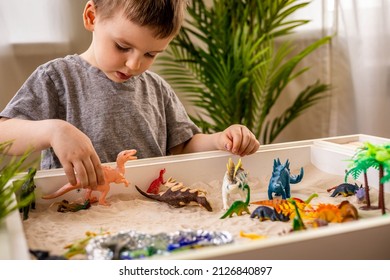 Cute baby boy playing sensory box dinosaur world kinetic sand table with carnivorous and herbivorous dinosaurs. Male kid enjoying early development game fine motor skills with Montessori material
