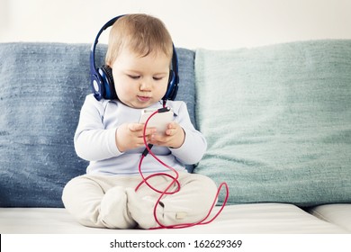 Cute Baby Boy Sitting On Sofa And Listening Music At Headphones While  Looking At Iphone.