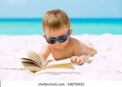 Cute baby boy with sunglasses lying on white sandy beach, reading the book and having his first tropical vacation. 