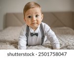 A cute baby boy wearing a bow tie and suspenders is seen crawling on a bed, displaying curiosity and innocence.