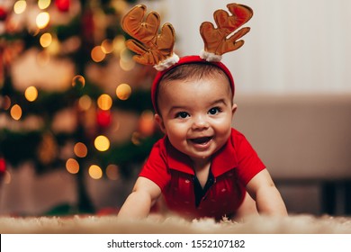 Cute Baby Boy Wearing Reindeer Antlers Crawling On Floor Over Christmas Lights. Looking At Camera. Holiday Season.