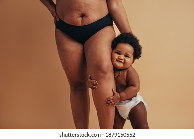 Cute Baby Clinging To Her Mother's Leg. Cropped Shot Of A Woman With Postpartum Body Standing With Her Baby.