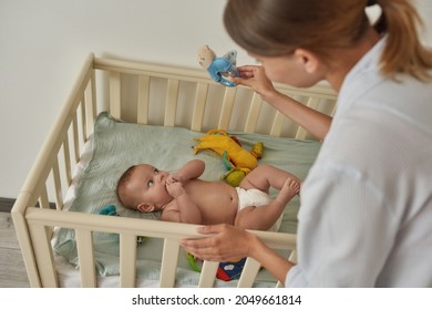 Cute Baby In Crib Watching Moving And Noisy Toy In Mother's Hand. Young Woman Holding Blue Rattle To Help Her Baby To Train New Skills. Children's Development Concept.