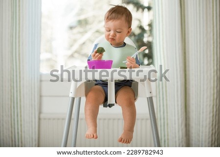 Cute baby eating with first solid food, infant sitting in high chair. Child doesn't want to eat, refuses eating, making faces. Cozy kitchen interior. Healthy food concept. 