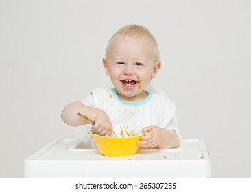 Cute Baby Eating In White High Chair Against White Background With A Yellow Bowl.  