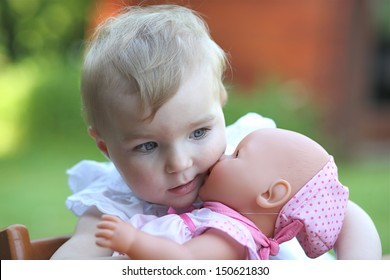 Cute baby girl with blond hair, blue eyes and white dress holding a baby doll