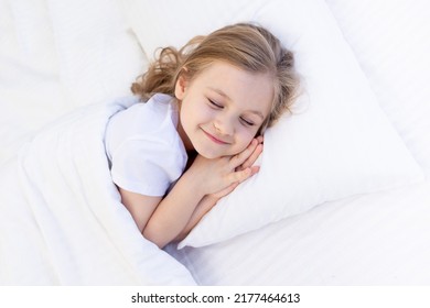A Cute Baby Girl Hugs A Pillow With Her Hands Folded Under Her Cheek And Sleeps On The Bed On A White Cotton Bed Under A Blanket, A Healthy Baby's Sleep At Night