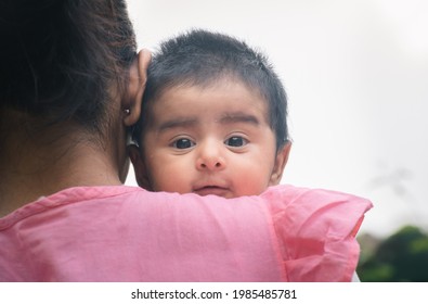 Cute Baby Girl Peeking Out From Mother's Shoulders With Eyes Wide Open, Looking At Camera. Two Months Old Sweet Daughter With Black Hair Hugging Close To The Warmth Of Her Mom.