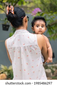 Cute Baby Girl Peeking Out From Mothers Shoulders With Eyes Wide Open, Looking At Camera. Five Months Old Sweet Daughter With Black Hair Hugging Close To The Warmth Of Her Mom.