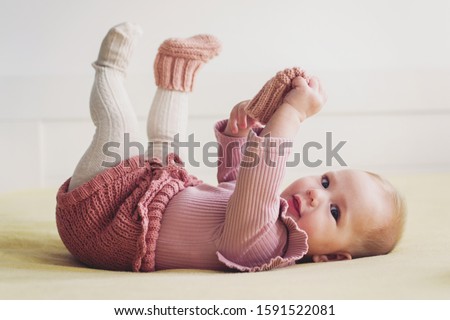 Cute baby girl playing with her sock, wearing dusty pink crochet bloomer and socks, smiling looking at camera, lying in bed
