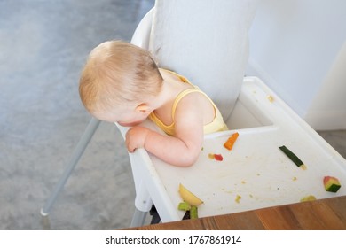 Cute baby girl sitting in highchair with fruit slices on tray and looking down at floor. High angle. Feeding process or child care concept