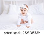 cute baby girl sitting on the bed in white clothes and with a bow on her head, funny little baby on a cotton bed at home and smiling, baby products concept