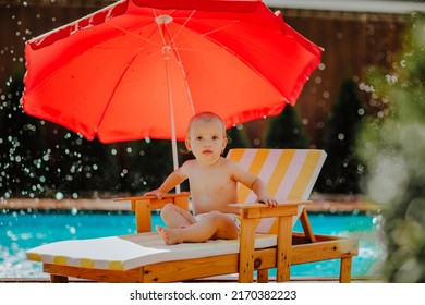 Cute Baby Girl Is Sitting On Small Stripped Beach Chair With Red Sun Umbrella Against The Pool With Splashing