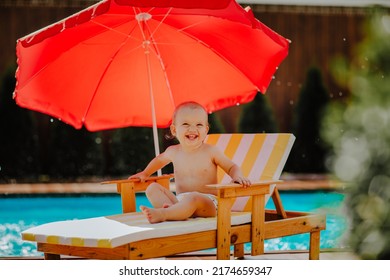 Cute Baby Girl Is Sitting On Small Stripped Beach Chair With Red Sun Umbrella Against The Pool With Splashing