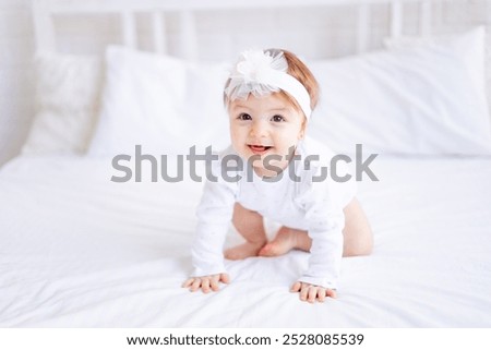 cute baby girl sitting on the bed in white clothes and with a bow on her head, funny little baby on a cotton bed at home and smiling, baby products concept