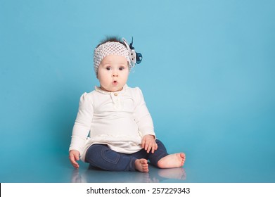Cute baby girl sitting in room over blue. Wearing trendy dress. Looking surprised at camera. Childhood.