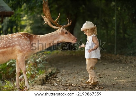 Cute baby girl, toddler, child feeding big brown deer with antlers in forest, park.Brave,cute caucasian blonde kid.Wild animal,wildlife and human concept