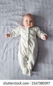 Cute Baby Girl In White Jumpsuit Lies On Grey Sheet Put On Bed At Home. Adorable Newborn Kid Spreads Hands To Sides In Bedroom Upper Closeup