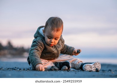 A cute baby in a green jacket explores pebbles on a sandy beach during sunrise in Victoria, Vancouver Island, BC. The serene morning setting captures curiosity and innocence. - Powered by Shutterstock