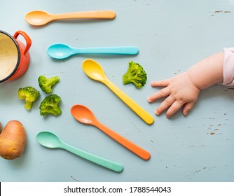 Cute Baby Hand Reaching For Spoon, Baby Food Concept, Vegetables, Spoons And Small Pot On The Table.