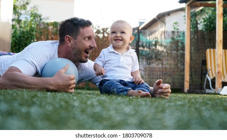 A Cute Baby And His Smiling Father Playing On A Grass With A Ball