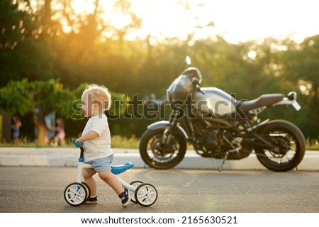 Cute baby learns to ride a balance bike. A small child tries to ride a bike in a sunny park with a big motorcycle in the background. The kid dreams of becoming a biker