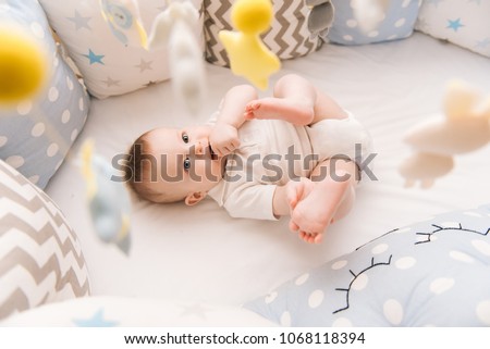 Cute baby lies in a white round bed. Light nursery for young children.  Toys for infant cot. Smiling child playing with mobile of felt and chewing his feet in sunny bedroom.