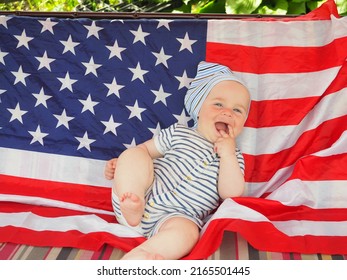 A Cute Baby Is Lying On The American Flag. A Happy Infant Plays Swinging On A Swing Against The Background Of The National Flag Of The United States Of America.