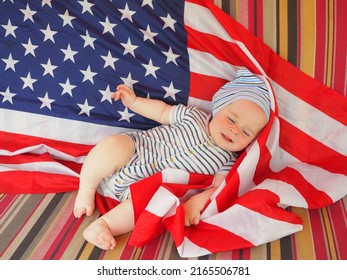 A Cute Baby Is Lying On The American Flag. A Happy Infant Plays Swinging On A Swing Against The Background Of The National Flag Of The United States Of America.