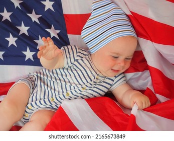 A Cute Baby Is Lying On The American Flag. A Happy Infant Plays Swinging On A Swing Against The Background Of The National Flag Of The United States Of America.