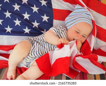 A Cute Baby Is Lying On The American Flag. A Happy Infant Plays Swinging On A Swing Against The Background Of The National Flag Of The United States Of America.