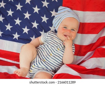 A Cute Baby Is Lying On The American Flag. A Happy Infant Plays Swinging On A Swing Against The Background Of The National Flag Of The United States Of America.