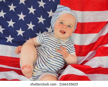 A Cute Baby Is Lying On The American Flag. A Happy Infant Plays Swinging On A Swing Against The Background Of The National Flag Of The United States Of America.