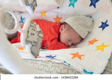 Cute Baby Newborn Boy Wearing Red Christmas Onesie And A Hat Sleeping On His Side In A Nest With Toys Hanging Over