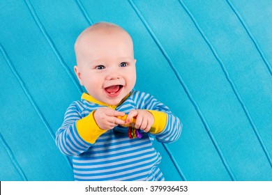 Cute Baby On Blue Knitted Blanket. Teething Infant Playing With Colorful Toy. Little Boy In Bed After Nap. Bedding And Textile For Nursery And Young Children. Kids Sleep Wear. Newborn Child At Home.