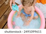 Cute baby playing in a colorful kiddie pool on a wooden deck. An adult pours water from a toy, creating a joyful and refreshing moment under the sun