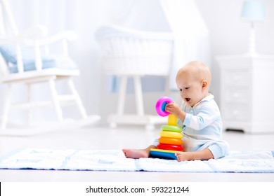 Cute Baby Playing With Colorful Rainbow Toy Pyramid Sitting On Play Mat In White Sunny Bedroom. Toys For Little Kids. Interior For Little Boy Nursery. Child With Educational Toy. Early Development.