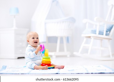 Cute Baby Playing With Colorful Rainbow Toy Pyramid Sitting On Play Mat In White Sunny Bedroom. Toys For Little Kids. Interior For Little Boy Nursery. Child With Educational Toy. Early Development.