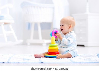 Cute Baby Playing With Colorful Rainbow Toy Pyramid Sitting On Play Mat In White Sunny Bedroom. Toys For Little Kids. Interior For Little Boy Nursery. Child With Educational Toy. Early Development.