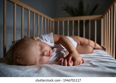 Cute Baby Sleeps At Night In A Cradle For Babies Without A Canopy And Bumpers. Safe Sleep In A Dark, Spacious Room With A Cot And A Flower. Interior Of The Bedroom And Children's Room