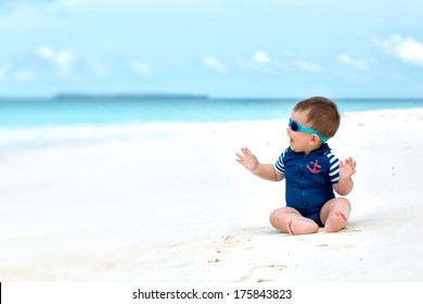 Cute Baby Smiling, Sitting On White Sandy Tropical Beach On Maldives, Having Vacation. With Diving Costume Or Swimwear, Sunglasses.