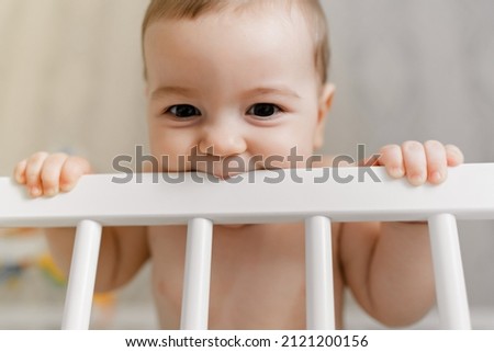 A cute baby is standing in the crib and biting the railing. Teeth eruption concept. Cute toddler kid biting on a crib, closeup portrait. Babys teething.