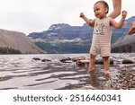 A cute baby takes first steps in the scenic landscape of Waterton, Alberta, Canada, assisted by a parent