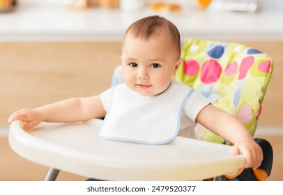 A cute baby wearing a white bib sits comfortably in a high chair in a well-lit kitchen. The baby looks curious and content while holding onto the edge of the high chair. - Powered by Shutterstock